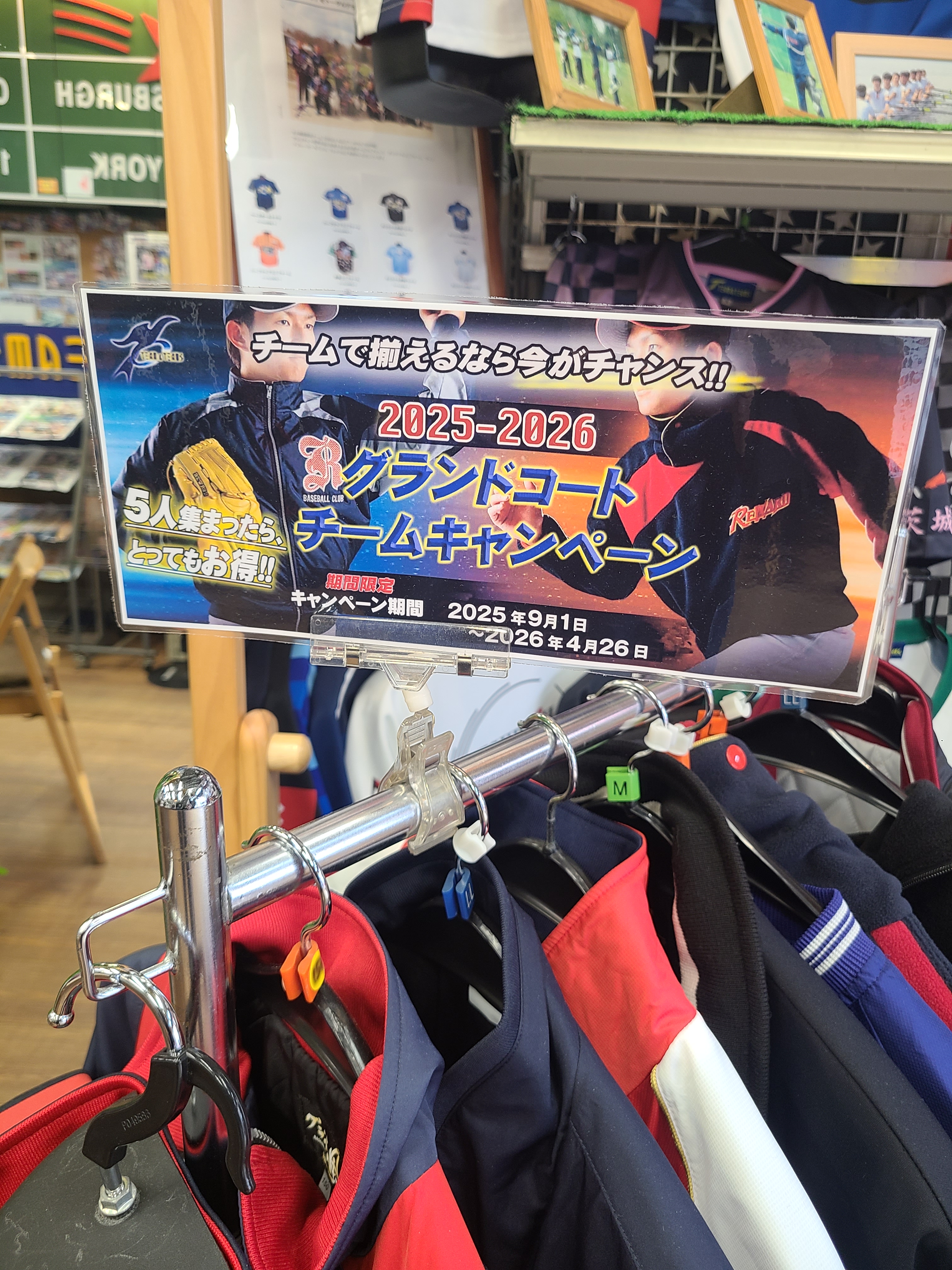 Clothing rack with red and navy jackets, plus a poster advertising 2025-2026 team campaign in Japanese posters in the background.
