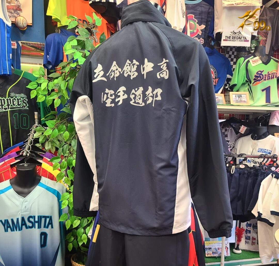 Navy windbreaker with white side panels and bold white kanji on the back, displayed in a sports shop among jerseys.