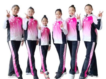 Seven young female athletes in matching pink gradient jackets and black pants, posing with peace signs against a white background.