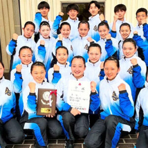 Group of young female athletes in white and blue tracksuits posing with fists raised, holding a plaque and a certificate in front of a doorway/studio.