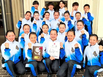 Group of young female athletes in white and blue tracksuits posing with fists raised, holding a plaque and a certificate in front of a doorway/studio.