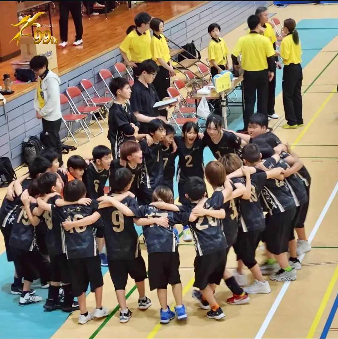 Young basketball players in black jerseys form a circle with arms around each other in a gym, preparing before a game as teammates and coaches stand nearby.